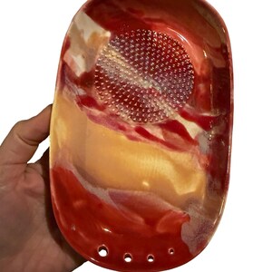 May include: An oval ceramic grater dish featuring a glossy glaze in shades of red, orange, and cream. The center has a raised, textured circular area for grating, and the bottom edge includes four small circular holes for drainage or hanging.