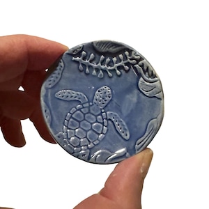 May include: A round, light blue ceramic piece featuring a detailed sea turtle design. The turtle is surrounded by embossed aquatic plants and wave-like patterns. The piece is held in a hand against a white background.