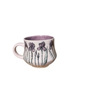 May include: A ceramic mug with a light pink exterior and a lavender interior. The mug is decorated with a black and purple floral design of irises. The handle is light pink.