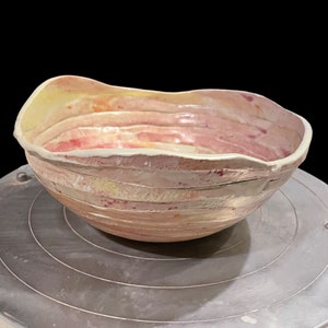 May include: A handmade ceramic bowl with a wavy rim and a layered, striped design in shades of pink, yellow, and white. The bowl is sitting on a pottery wheel.