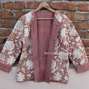 Puede incluir: Una chaqueta acolchada marrón con estampado floral blanco. La chaqueta tiene un forro a rayas rojo y azul y un cierre de botón. La chaqueta está colgada de una percha de madera contra una pared de ladrillo.