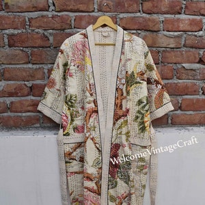 Op de afbeelding: Een crèmekleurige badjas met een bloemenprint en zwarte bies. De badjas heeft een strikceintuur en lange mouwen. De badjas hangt aan een houten hanger tegen een bakstenen muur.