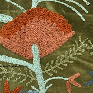 May include: An olive green velvet fabric featuring intricate punch needle embroidery. The design displays a large rust-colored flower with light blue stems and leaves, alongside smaller floral elements in blue and orange. Text reads "elcomeVintageCrafts".