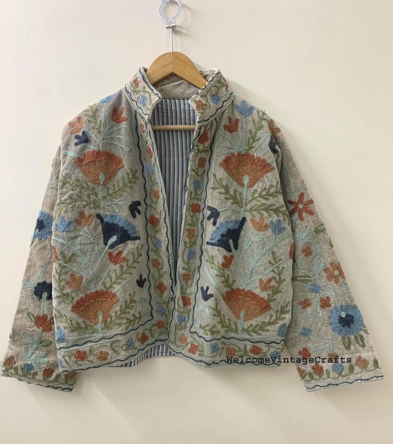 May include: A light beige jacket featuring intricate floral embroidery in shades of orange, blue, and green. The garment has a high collar, long sleeves, and an open front. The interior lining displays a blue and white vertical striped pattern.