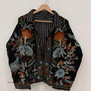 May include: A black jacket featuring long sleeves, a collar, and an open front. The fabric is decorated with intricate floral embroidery in shades of orange, light blue, and green. The interior lining displays a black and white vertical stripe pattern.
