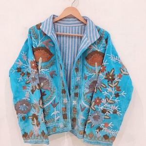 May include: A bright turquoise jacket featuring intricate floral embroidery in shades of brown, blue, and white. The garment has long sleeves, a collared neckline, and an interior lining with a blue and white vertical pinstripe pattern.