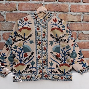 May include: A beige embroidered jacket with a floral pattern in shades of blue, green, red, and black. The jacket has a mandarin collar and a button closure.