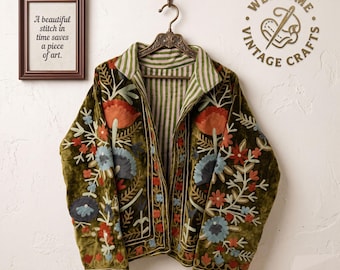May include: An olive green velvet jacket featuring intricate floral embroidery in shades of orange, blue, and red. The jacket has a collared neckline and an interior lining with a green and white vertical stripe pattern, hanging on a metal hanger.