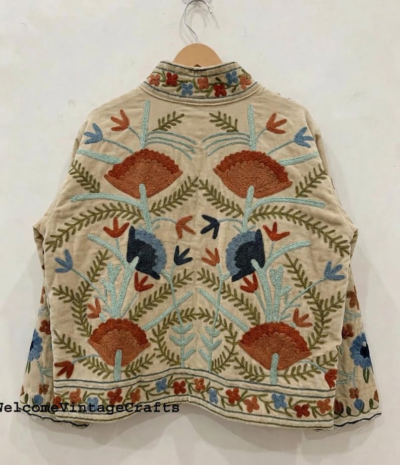 May include: A beige jacket featuring intricate floral embroidery in shades of rust orange, light blue, and dark navy. The design includes symmetrical botanical patterns across the back and sleeves, with a high collar and a straight hemline.