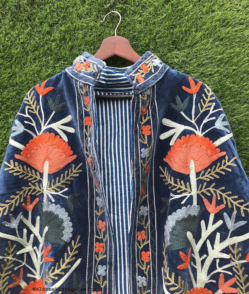 May include: A dark blue velvet jacket featuring vibrant orange, light blue, and grey floral embroidery. The garment has a high collar and an interior lining with a white and blue vertical stripe pattern. The text WelcomeVintageCrafts is visible.
