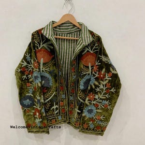 May include: Olive green velvet jacket with colorful floral embroidery. The jacket features a striped collar and lining, and is displayed on a wooden hanger. The embroidery includes orange, blue, and red flowers.