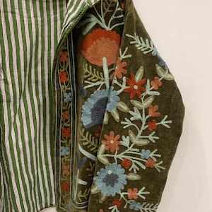 May include: An olive green jacket featuring intricate floral embroidery in shades of orange, light blue, and dark blue. The interior lining displays a green and white vertical striped pattern. The text WelcomeVintageCrafts is visible at the bottom.