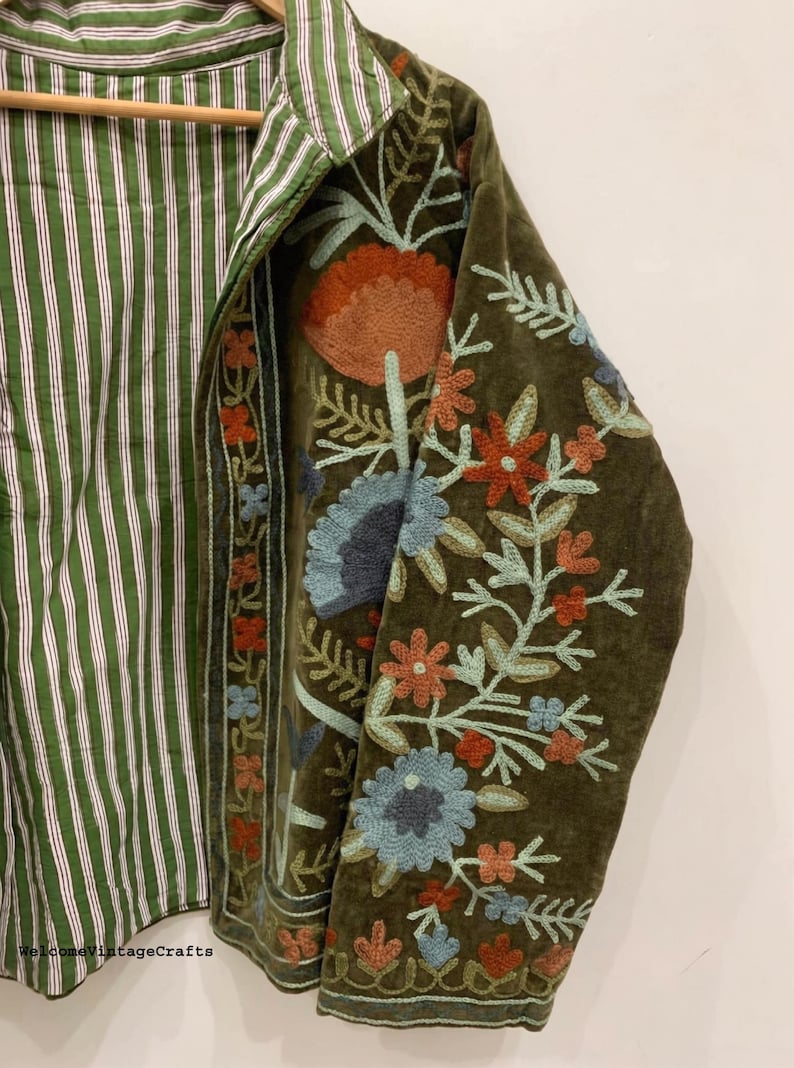 May include: An olive green jacket featuring intricate floral embroidery in shades of orange, light blue, and dark blue. The interior lining displays a green and white vertical striped pattern. The text WelcomeVintageCrafts is visible at the bottom.
