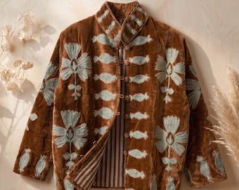 Puede incluir: Una chaqueta de terciopelo marrón con motivos florales bordados en azul claro y patrones decorativos horizontales. La prenda tiene cuello alto, mangas largas y cierre frontal con pequeños botones marrones, dejando ver un forro interior a rayas.