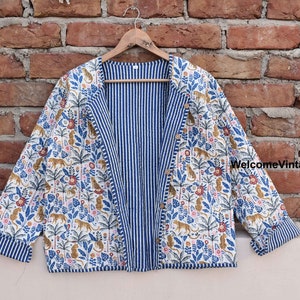May include: A reversible jacket with a white background, featuring a jungle print with leopards, palm trees, and flowers. The reverse side has blue and white vertical stripes. The jacket is hanging on a wooden hanger.