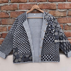 Peut inclure: Une veste matelassée noire et blanche avec une doublure rayée. La veste présente un design patchwork avec différents motifs, notamment des cercles, des points et des motifs floraux. La veste est ouverte et suspendue à un cintre en bois.