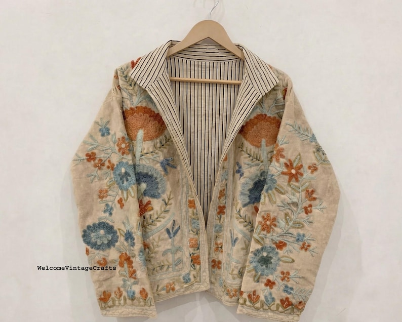 May include: A beige jacket featuring intricate floral embroidery in shades of orange, blue, and green. The garment has a notched collar and an interior lining with a black and white vertical pinstripe pattern. The text WelcomeVintageCrafts is visible.