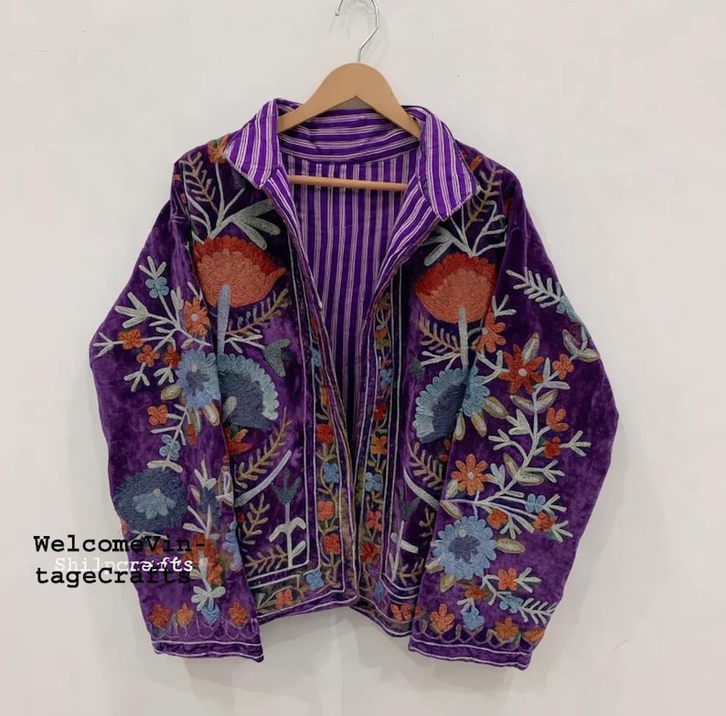 May include: A purple velvet jacket featuring intricate floral embroidery in shades of orange, blue, and white. The jacket has a collared neckline, long sleeves, and an interior lining with a purple and white vertical stripe pattern.