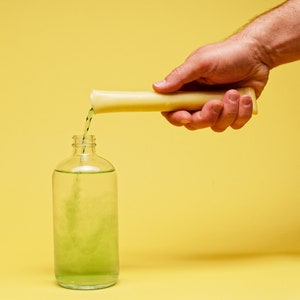 Puede incluir: Una mano vierte líquido verde de un tubo amarillo en una botella de vidrio transparente que contiene líquido verde.