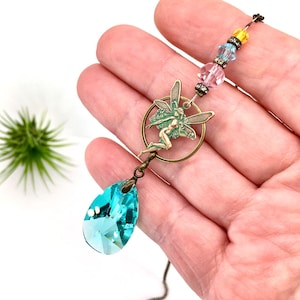 May include: A handcrafted necklace featuring a teal teardrop crystal pendant. The pendant is connected to a bronze-toned fairy charm and adorned with pink, blue, and yellow beads. The necklace is held in a hand against a white background.