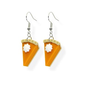 May include: A pair of dangle earrings shaped like pumpkin pie slices. Each earring features an orange pie filling, a tan crust, and a dollop of white whipped cream. The earrings hang from silver-colored fish hook ear wires.