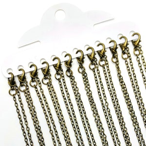 Pode incluir: Um conjunto de 10 correntes douradas com fechos de lagosta, apresentado em um cartão branco.