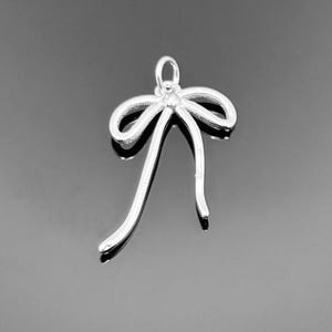 Puede incluir: Un colgante plateado con forma de lazo con un bucle en la parte superior para colgar. El lazo tiene una forma clásica con dos colas largas y curvas. El colgante probablemente está hecho de metal y tiene un acabado brillante. El fondo es una superficie oscura y reflectante.