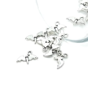 May include: Silver-colored bat charms scattered on a white surface. The charms have a detailed design, including wings and a small loop for attaching to jewelry or other crafts. The charms are small, with a detailed design.