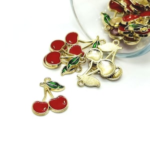 May include: A collection of gold-toned metal cherry charms with red enamel fruit and green enamel leaves. Several charms are spilling out of a clear glass container, while one individual cherry charm lies flat on the surface in the foreground.