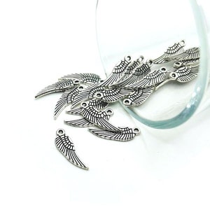 May include: A pile of silver metal angel wing charms, scattered on a white surface. The charms are detailed with a black outline and have two holes for attaching to jewelry.