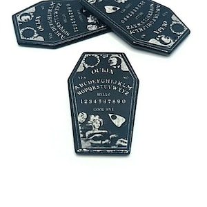 Puede incluir: Un tablero de Ouija negro en forma de ata&uacute;d con una planchette y letras plateadas. El tablero presenta las letras del alfabeto, los n&uacute;meros del 0 al 9 y las palabras "s&iacute;", "no", "hola" y "adi&oacute;s".
