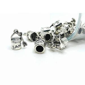 May include: A collection of silver charm pendants shaped like miniature buckets and spades, with the word "SAND" engraved on each. The charms are scattered on a white surface.
