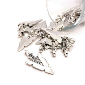 May include: A pile of silver arrowhead charms, some with a loop at the top, others with a loop at the bottom. The charms are scattered on a white surface.