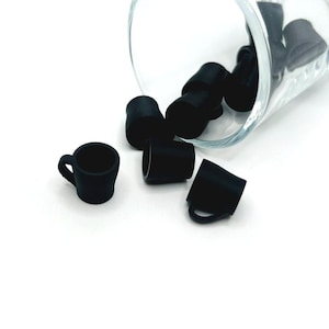 May include: Black miniature coffee cup-shaped bottle stoppers scattered on a white surface, with some spilling from a clear glass. The stoppers have a small handle and a cylindrical body. The image is well-lit, highlighting the details of the stoppers.