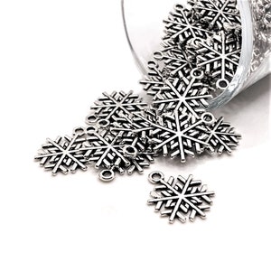 Op de afbeelding: Een stapel zilveren sneeuwvlokcharms met een lus aan de bovenkant. De charms zijn verspreid over een witte achtergrond.