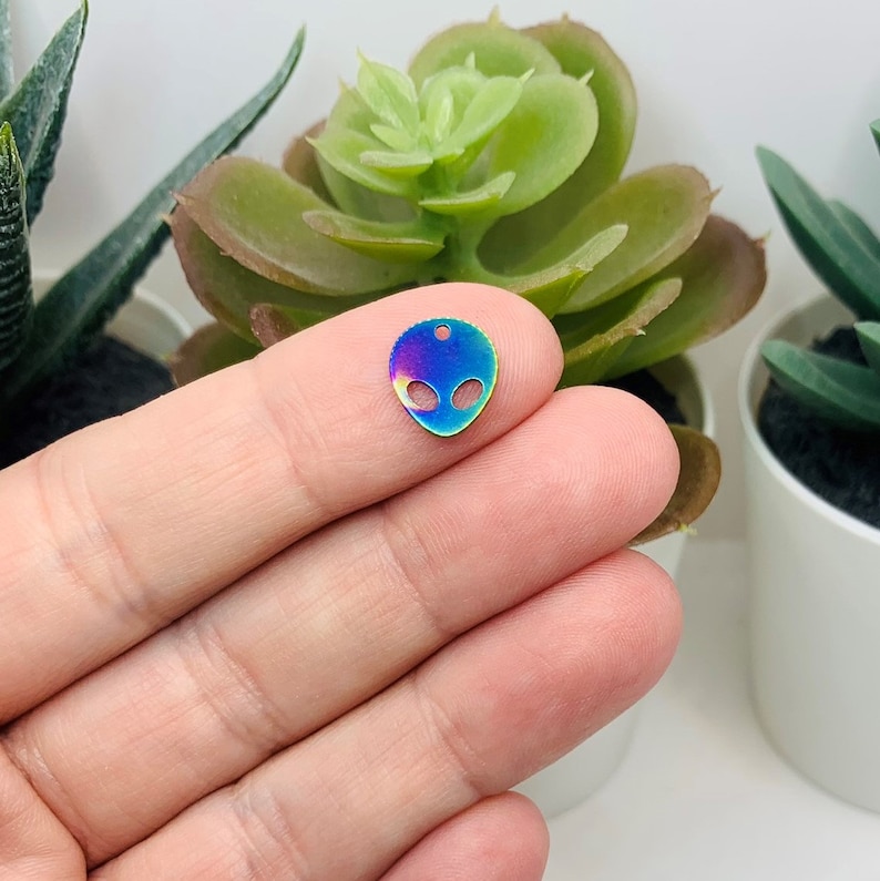 K&ouml;nnte beinhalten: Ein kleiner, schillernder Alien-Anh&auml;nger mit einem Loch zum Aufh&auml;ngen. Der Anh&auml;nger ist aus Metall und hat eine Regenbogen-Oberfl&auml;che.