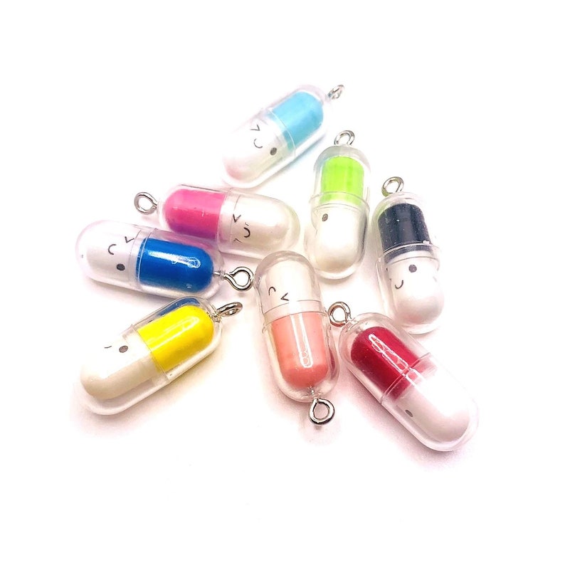 Op de afbeelding: Een verzameling van tien kleurrijke pil-charms met vrolijke gezichten. De charms zijn gemaakt van plastic en hebben een heldere, transparante behuizing. Elke charm heeft een andere kleur en een andere vrolijke gezichtsuitdrukking.