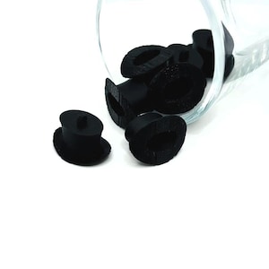 May include: Black rubber stoppers, some inside a clear glass container, others scattered on a white surface. The stoppers have a mushroom-like shape, with a flat base and a cylindrical top. The stoppers are likely for bottles or containers.