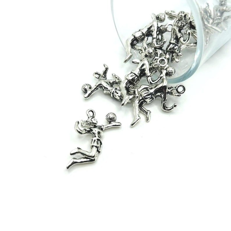 May include: A silver charm of a volleyball player spiking the ball. The charm is part of a group of charms spilling out of a glass jar.