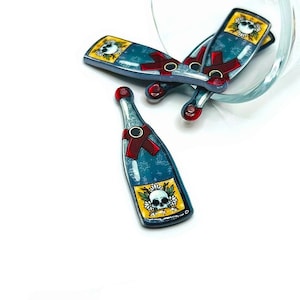 May include: Four blue glass bottle charms with a skull and floral design on the label. The bottles have red ribbon accents and are arranged on a white surface.