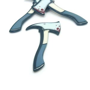 May include: Several small, stylized axe-shaped pins. The axes have light blue handles, white blades with red blood splatter details, and a dark blue outline. The pins are arranged on a white background.