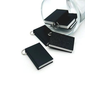 Puede incluir: Llaveros de libros negros en miniatura con anillas plateadas. Varios de los pequeños libros están esparcidos alrededor de un vaso transparente. Los libros tienen páginas blancas y un acabado liso y mate. Un libro tiene una cubierta texturizada.