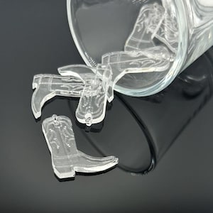 Op de afbeelding: Een groep transparante cowboylaarscharms van plastic valt uit een glazen pot op een zwart oppervlak. De charms zijn gedetailleerd met stiksels en een puntige neus.