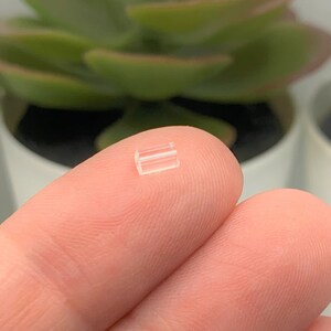 May include: A clear plastic earring back, designed to hold an earring in place. The back is small and rectangular, with a slight curve to fit the ear.