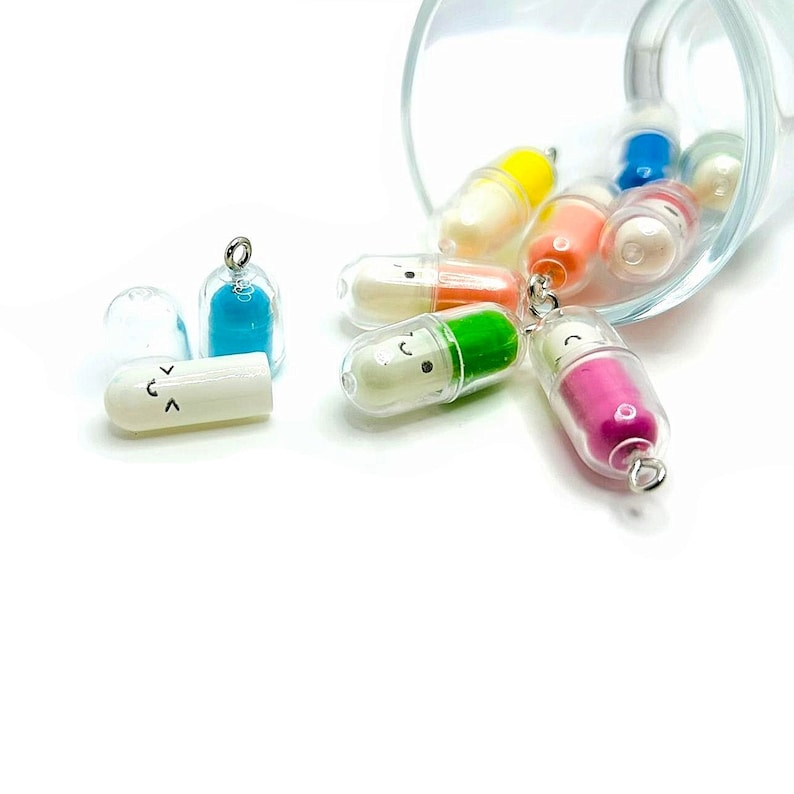 Op de afbeelding: Een glazen pot gevuld met kleurrijke capsules met vrolijke gezichten. De capsules vallen uit de pot op een wit oppervlak.