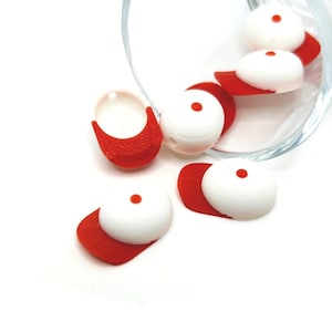 May include: White and red miniature baseball cap-shaped beads with a small red dot on top. The beads are scattered around a clear glass container, with some caps lying flat and others upright. The caps have a rounded top and a curved brim.