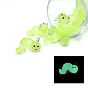 May include: Several translucent green caterpillar charms with smiling faces and silver-colored jump rings. Some are scattered, while others spill from a clear glass container. A smaller image shows a glowing caterpillar.