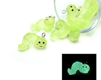 May include: A collection of translucent green caterpillar charms with smiling faces and silver jump rings. Some are scattered, others spill from a clear glass container. A smaller image shows a glowing caterpillar.
