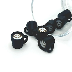 May include: A collection of small, black coffee cup charms with white latte art designs. The charms are arranged near a clear glass, showcasing the miniature details of each cup.
