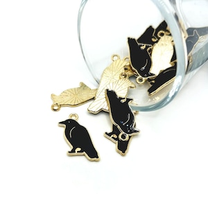 May include: Black and gold raven charms with a glossy finish. The charms are in various poses, some standing and others with their wings spread. A clear glass is partially visible in the background.